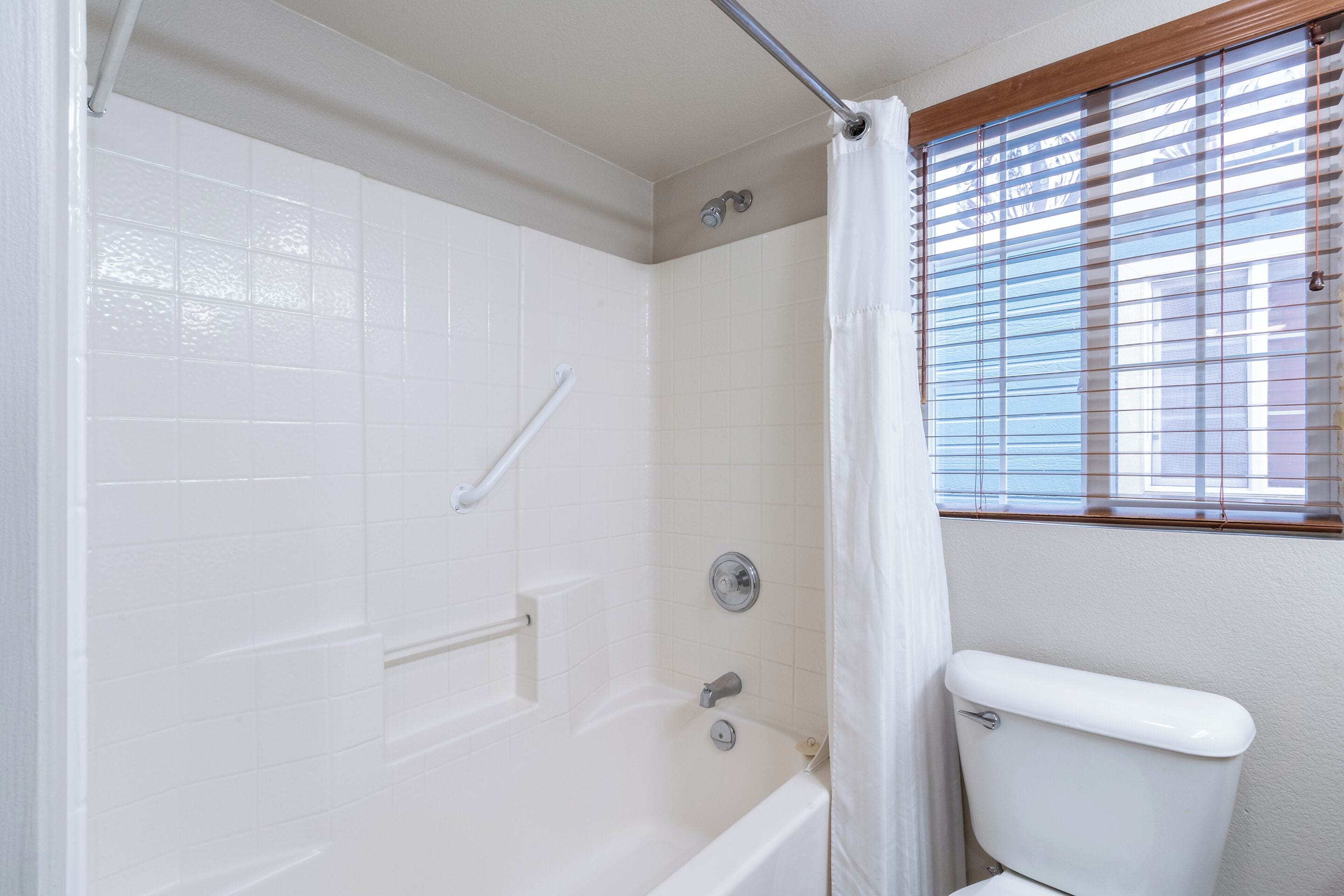 Ein Badezimmer mit weißen Fliesen, einer Badewanne, einer Toilette und einem Fenster mit Jalousien.
