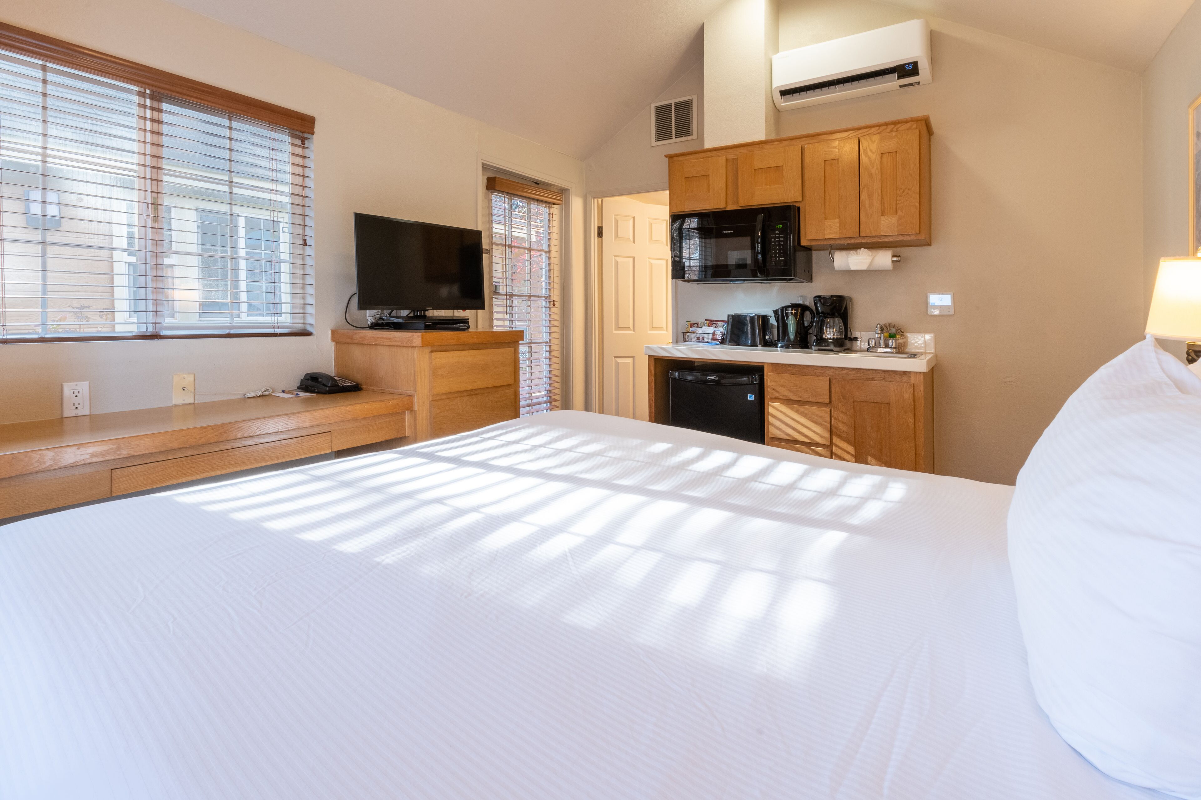 A hotel room with a large bed, a TV, a kitchenette with a microwave, and a window with blinds.