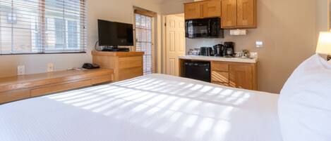 A hotel room with a large bed, a TV, a kitchenette with a microwave, and a window with blinds.