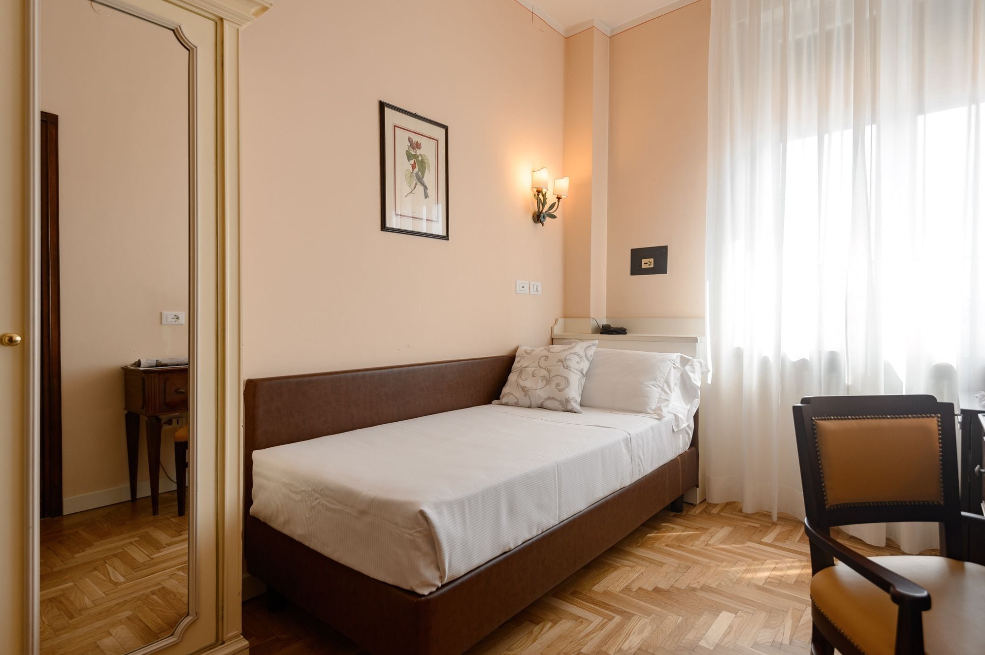 A bedroom with a bed, a chair, a nightstand, a wall-mounted lamp, and a framed picture.