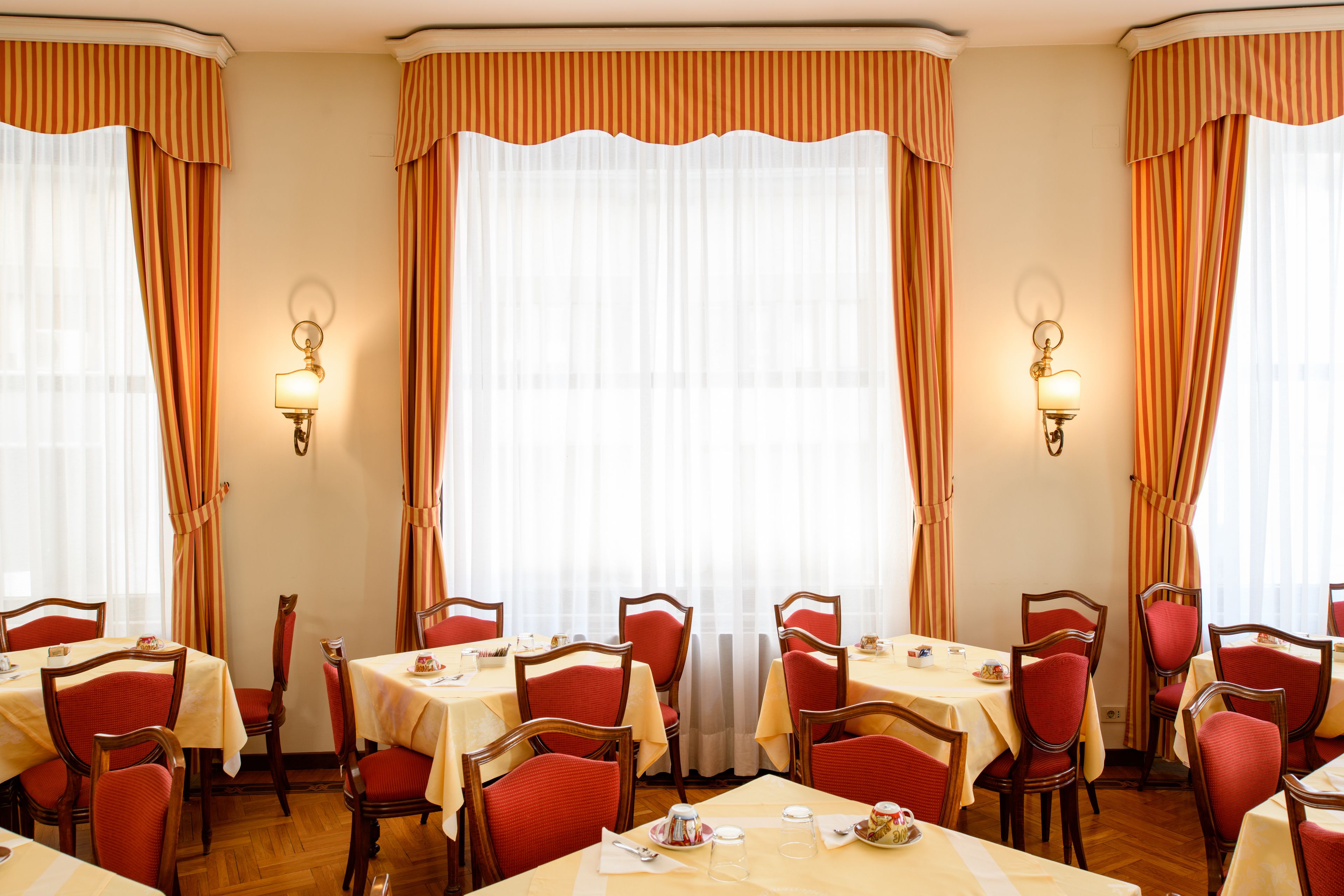 A dining area with tables and chairs, large windows with curtains, and wall-mounted lamps.