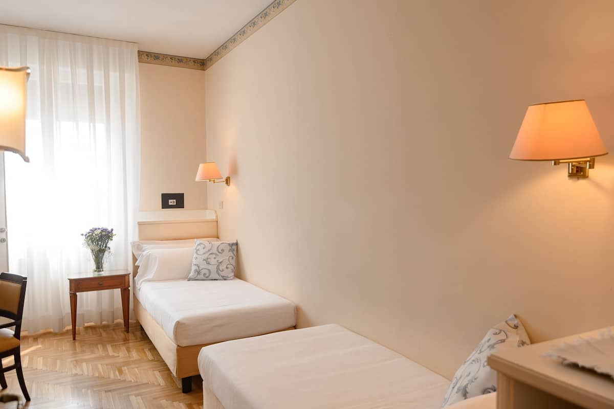 A hotel room with two single beds, a small table, a chair, and a lamp.