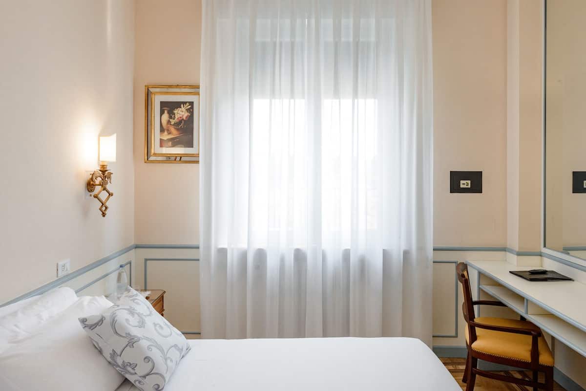 A hotel room with a bed, a desk, a chair, a window with sheer curtains, a wall-mounted light fixture, and a framed picture.