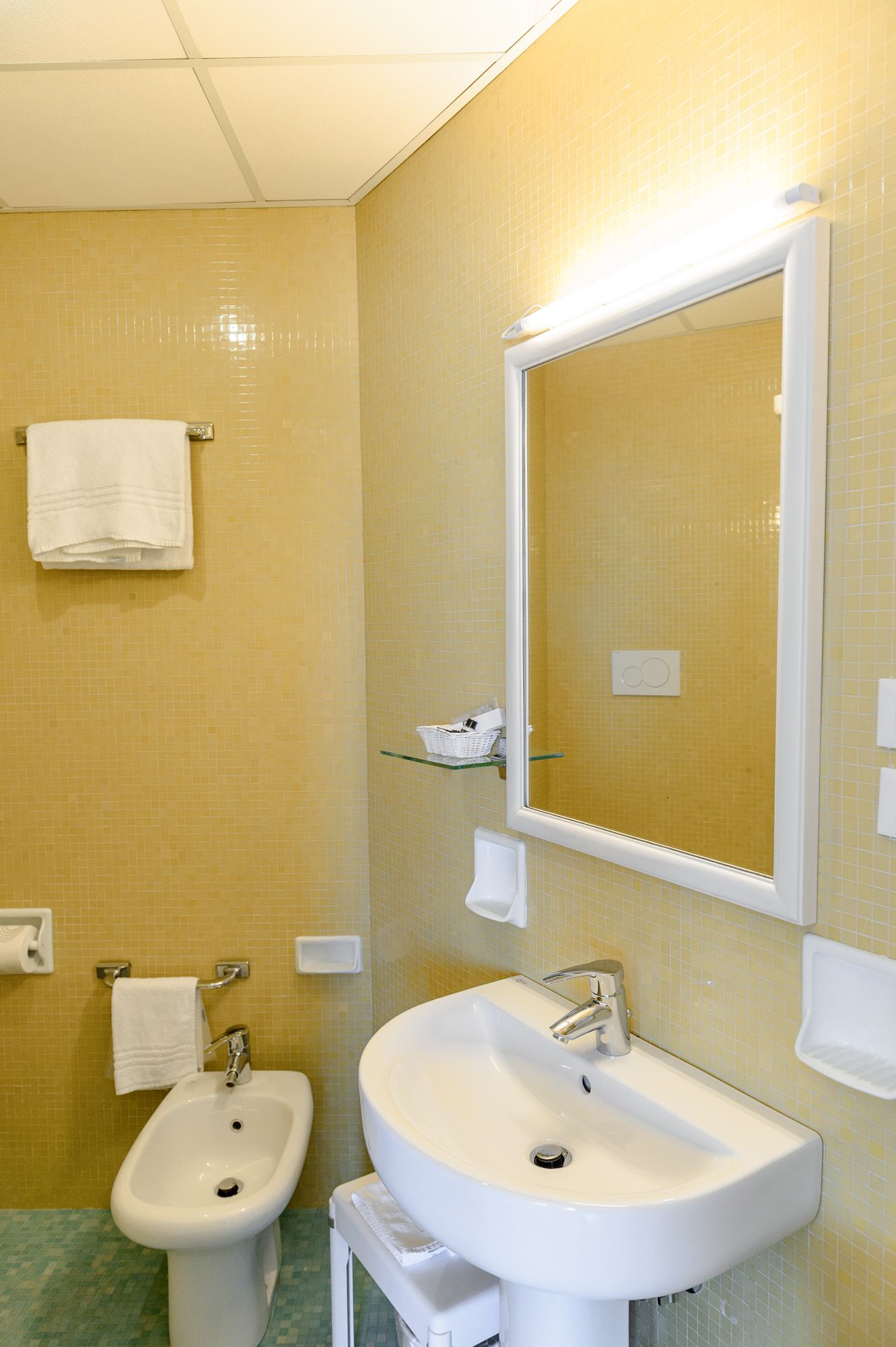 A bathroom with a yellow tiled wall, a white sink, a bidet, and a mirror.