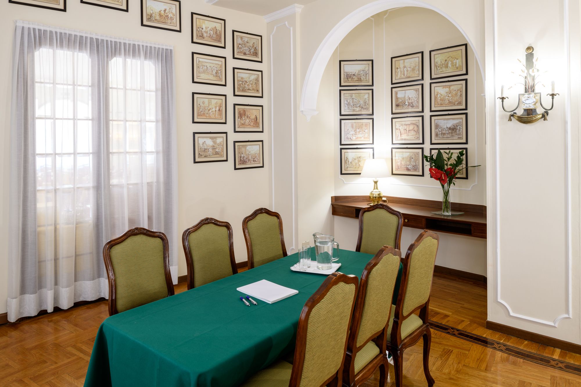 A conference room with a green table, six chairs, framed pictures on the walls, a desk with a lamp, and a vase with flowers.