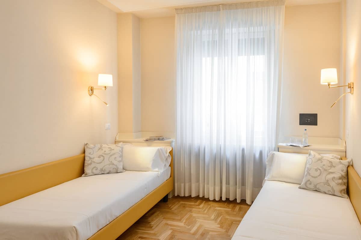 A hotel room with two single beds, a large window with sheer curtains, and wall-mounted lamps.
