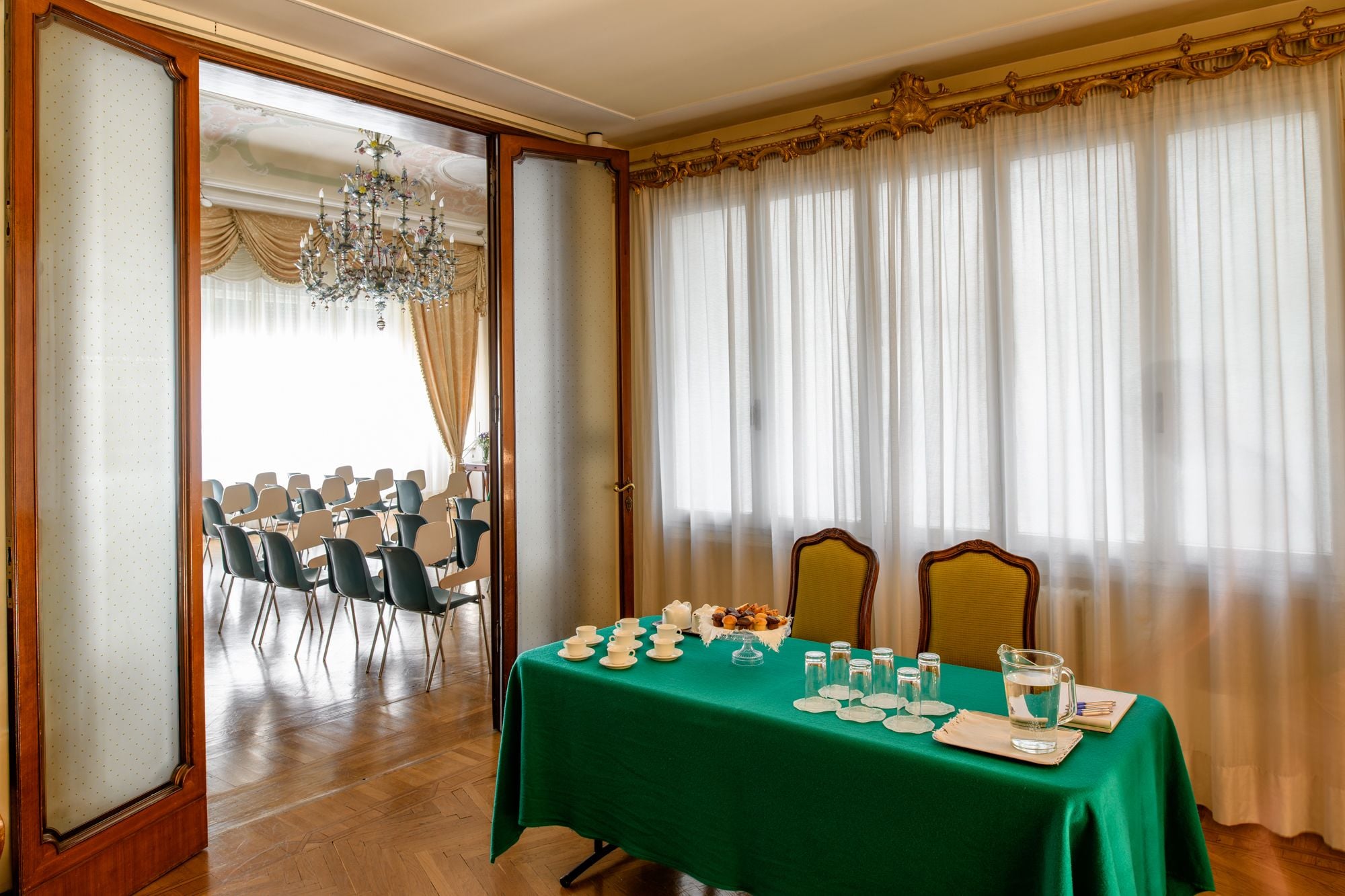A room with a green table set for a meeting, two chairs, a chandelier, and a view of a conference room with chairs.