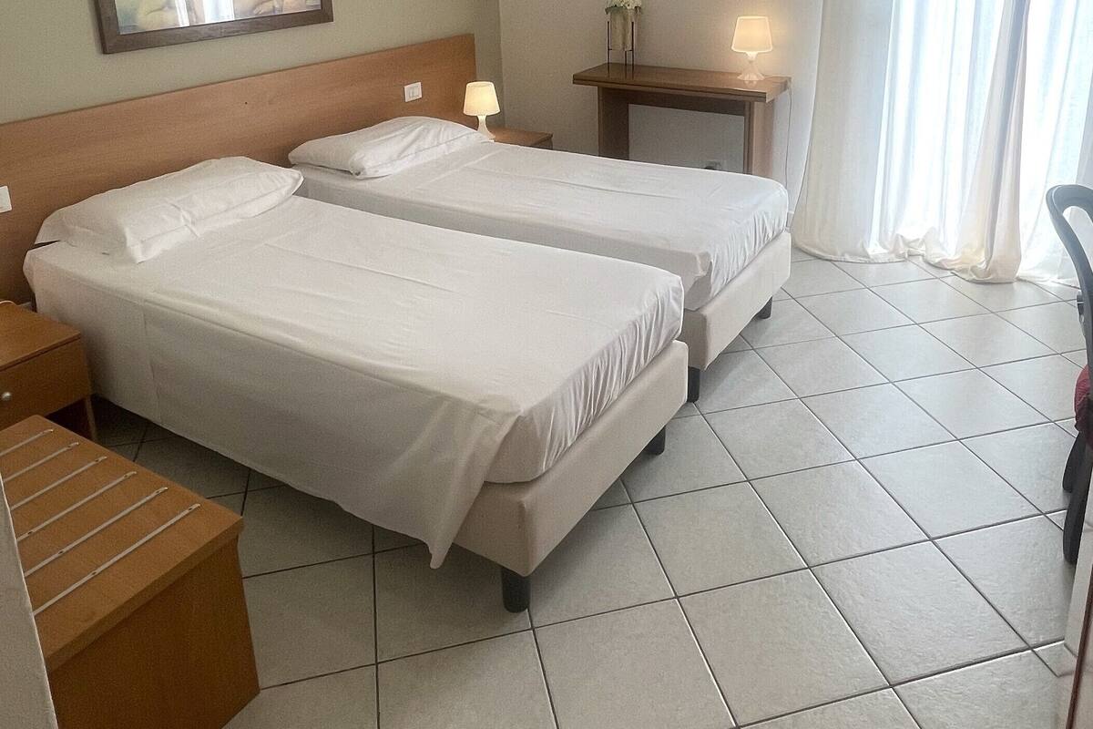 A hotel room with two beds, a nightstand, a small table with a lamp, and a window with curtains.