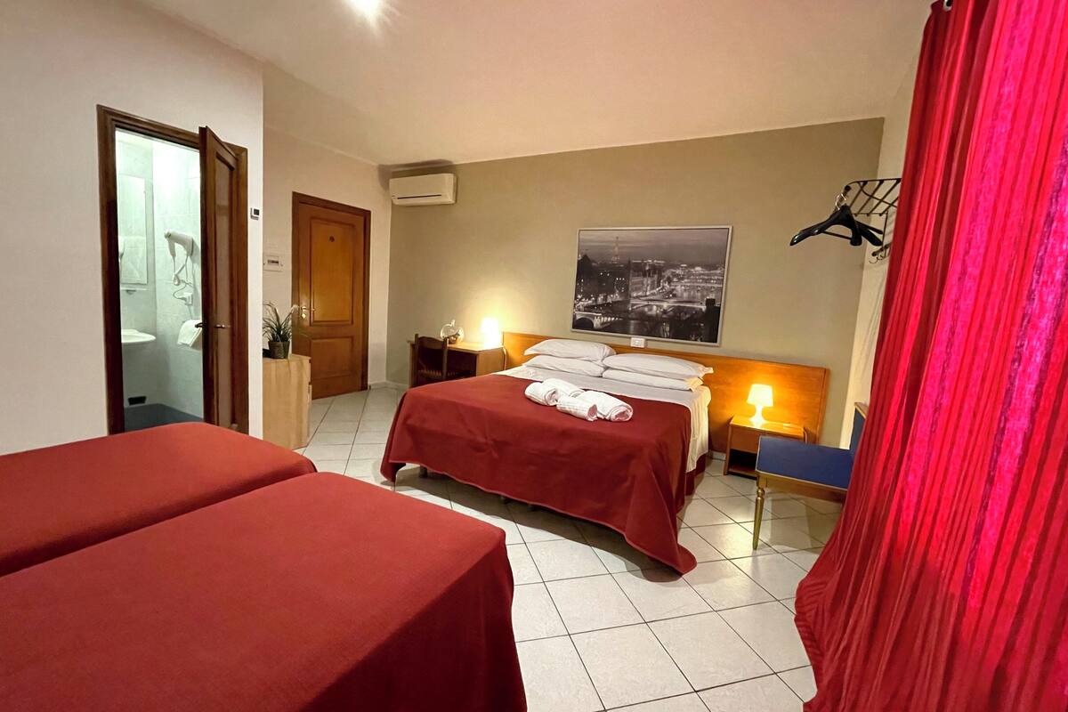 A hotel room with two beds, a bathroom, a nightstand with a lamp, and a cityscape painting on the wall.