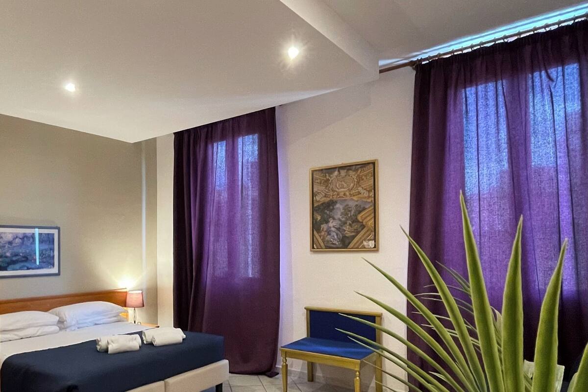 A hotel room with a bed, a chair, a desk, a lamp, a painting, and a large plant.