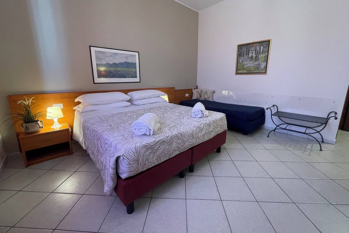 A hotel room with a large bed, a smaller bed, a bench, a nightstand with a lamp, and a wall-mounted painting.