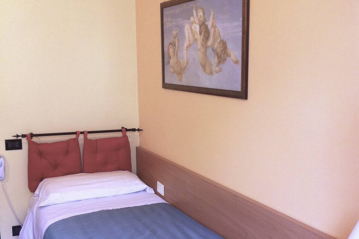 A single bed with a blue bedspread, a wooden headboard, a painting of cherubs on the wall, a bedside table with a remote control, and a telephone on the wall.
