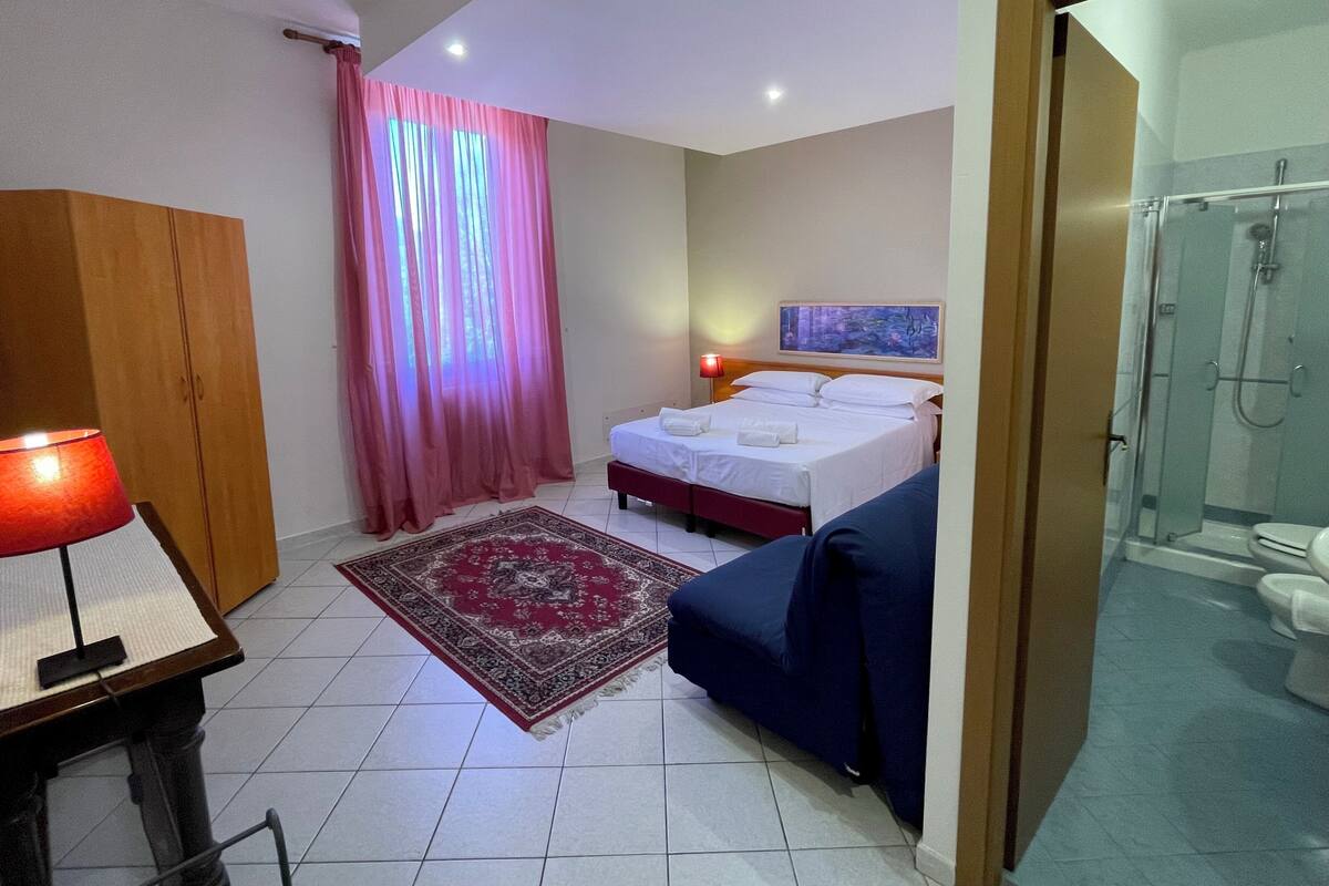 A hotel room with a bed, a desk with a lamp, a blue sofa, a shower area, and a painting on the wall.