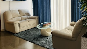 A modern living room with a beige sofa, a glass coffee table, and a TV mounted on the wall displaying a travel program. - Hotel San Carlo (Mestre)