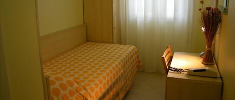 A small, single-bed room with a television on the wall, a desk with a vase of flowers, and a chair.