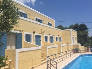 A building with a pool, featuring a staircase leading to a balcony with blue shutters, and a textured yellow exterior. - Esperides Maisonettes (Aegina)