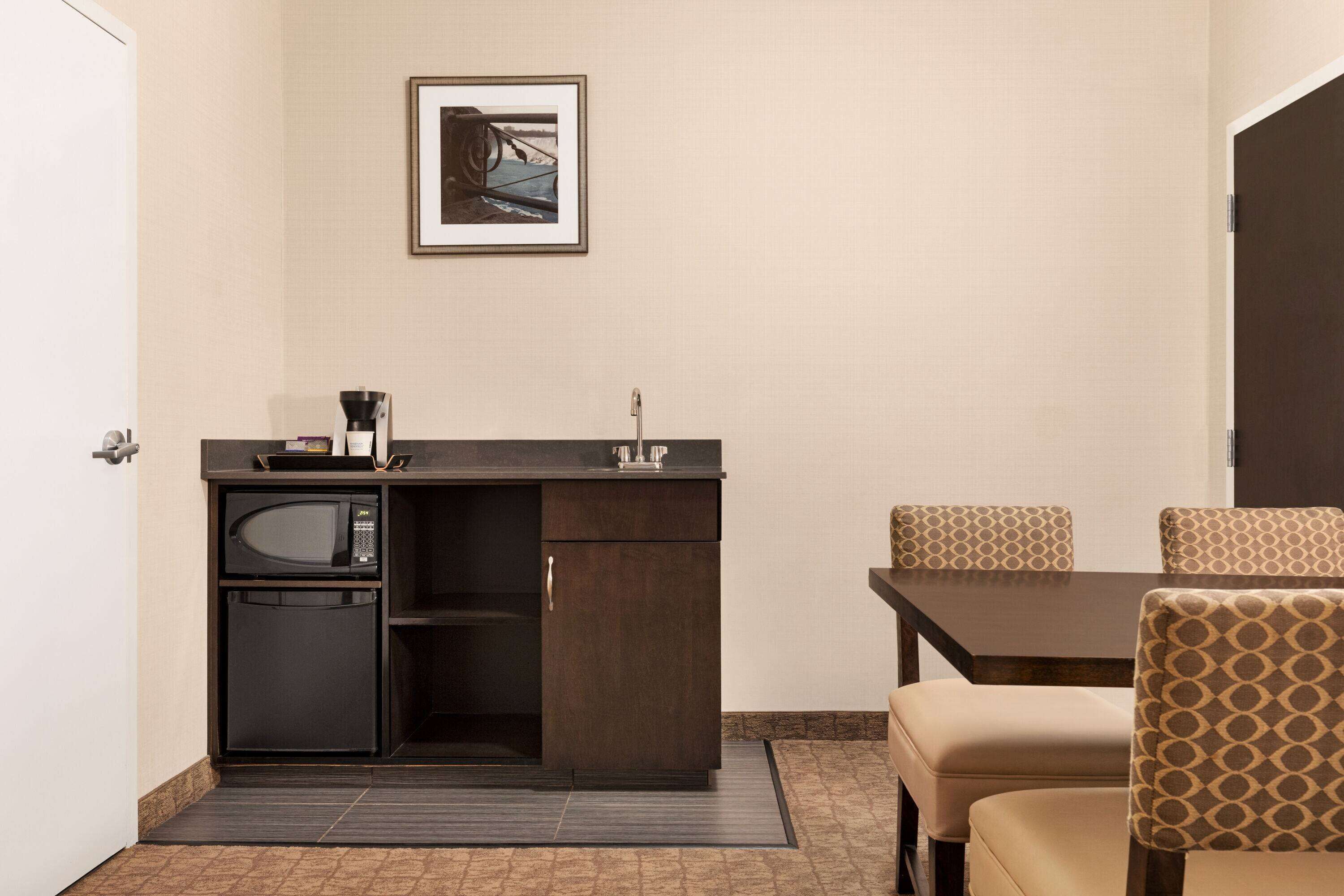A compact kitchenette with a microwave, refrigerator, and coffee maker. There is a framed picture on the wall and a dining table with patterned chairs.