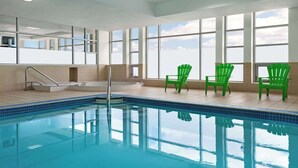 Una piscina interior con agua azul cristalina, rodeada de amplias ventanas y sillas verdes.