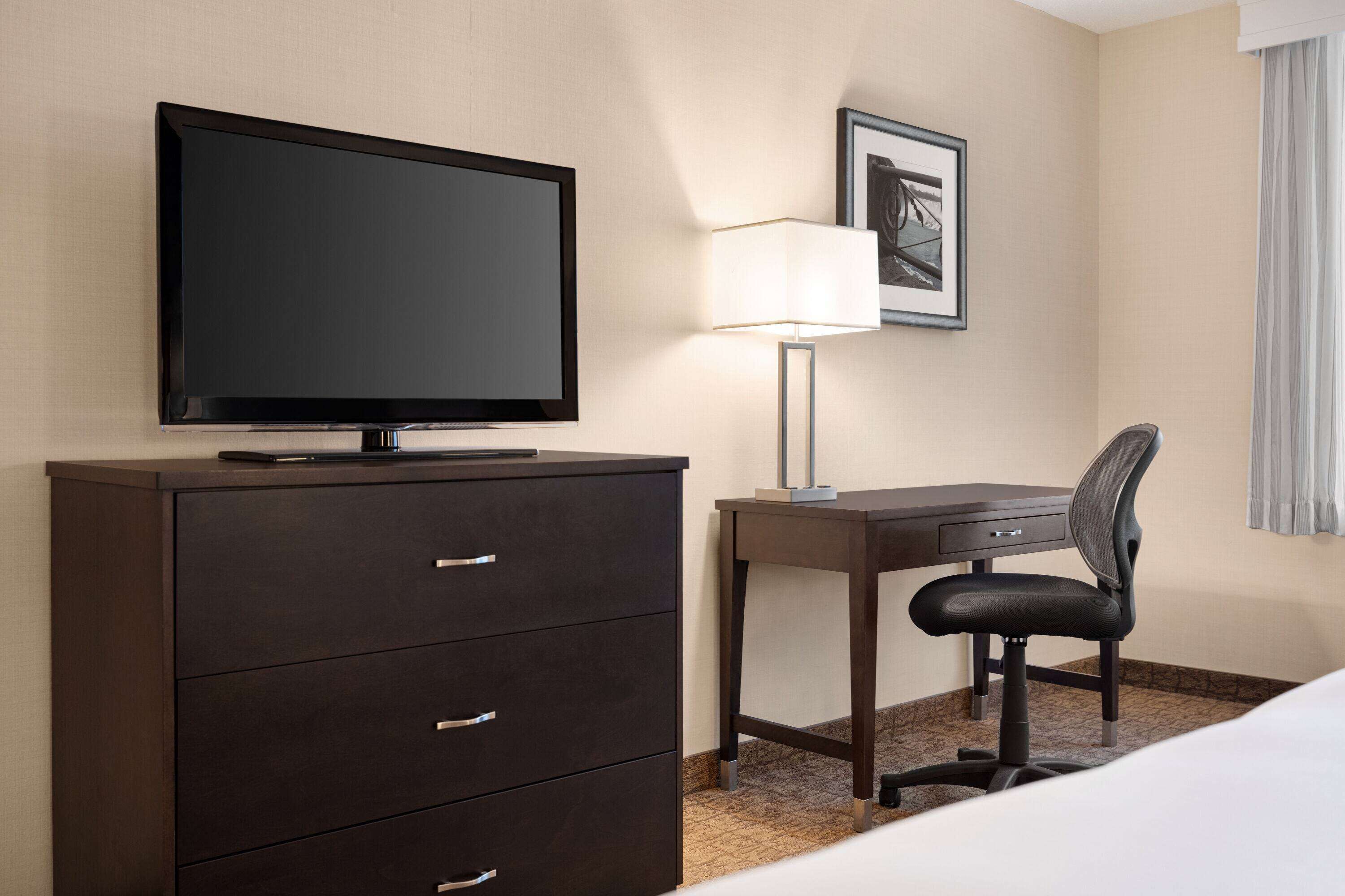 Habitación de hotel con televisor de pantalla plana sobre una cómoda de madera oscura, un escritorio con una silla, una lámpara y un cuadro en la pared.