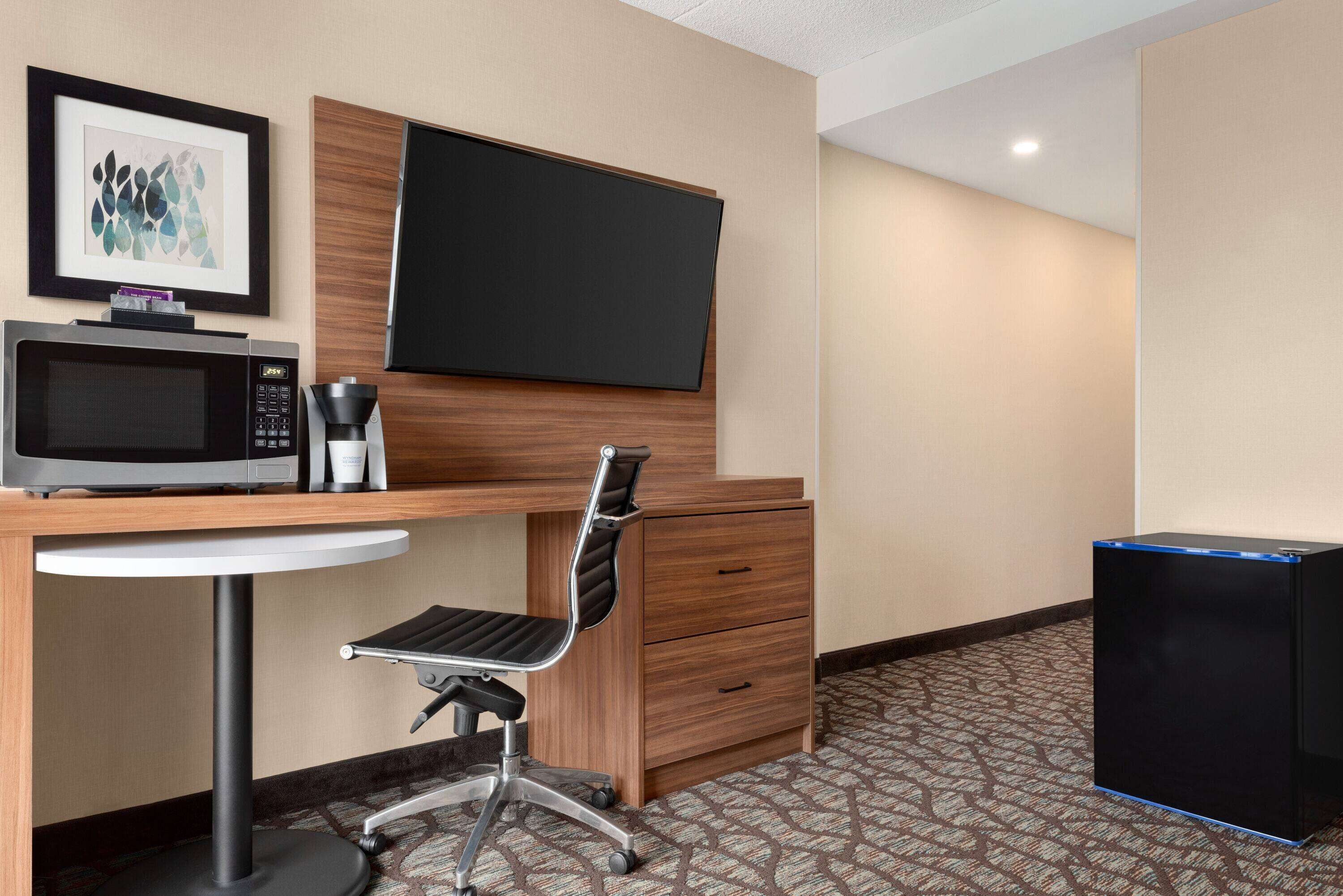 A hotel room with a desk, a chair, a microwave, a coffee maker, a flat-screen TV, and a trash bin.