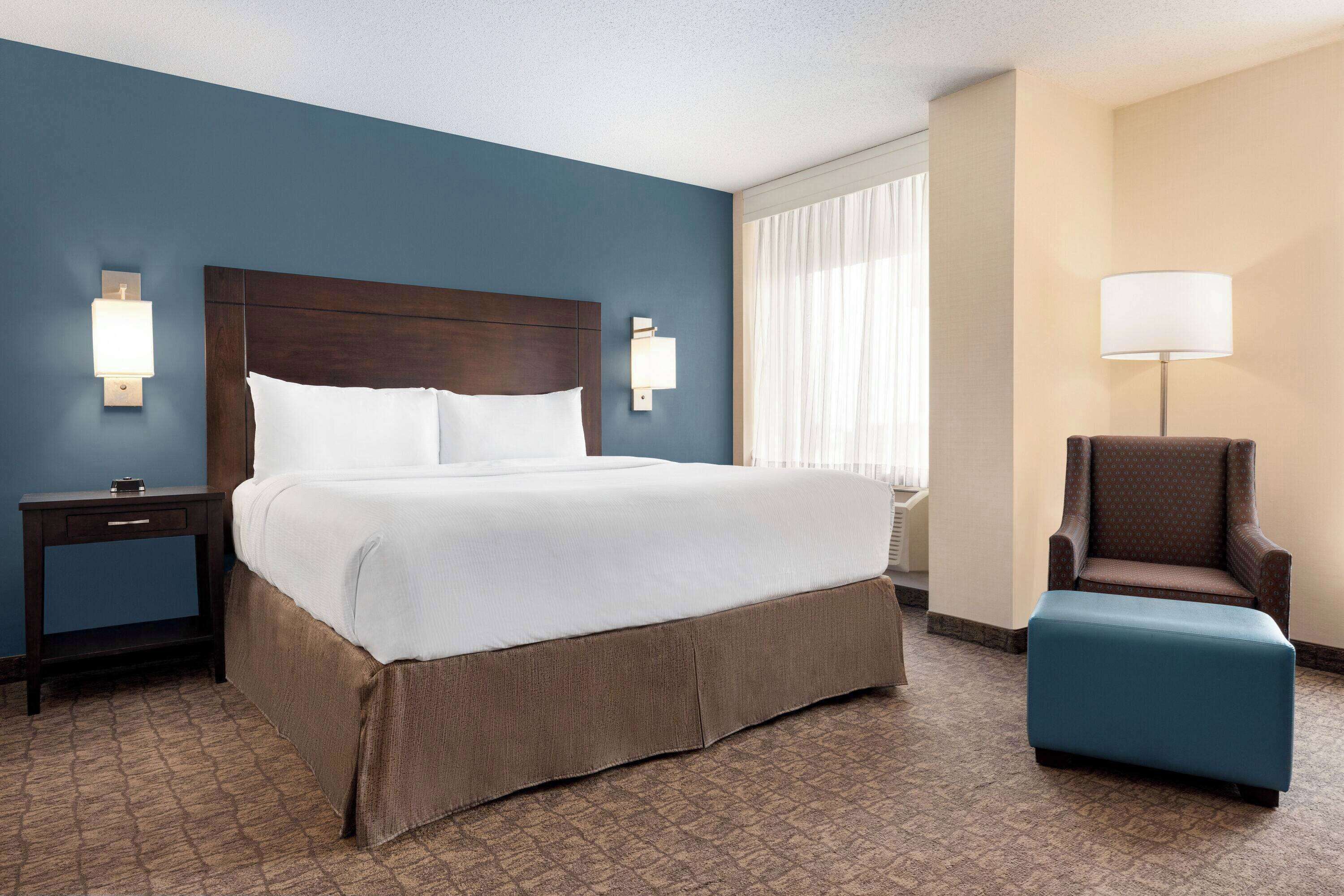 A hotel room with a large bed, a nightstand, a chair, and a lamp.