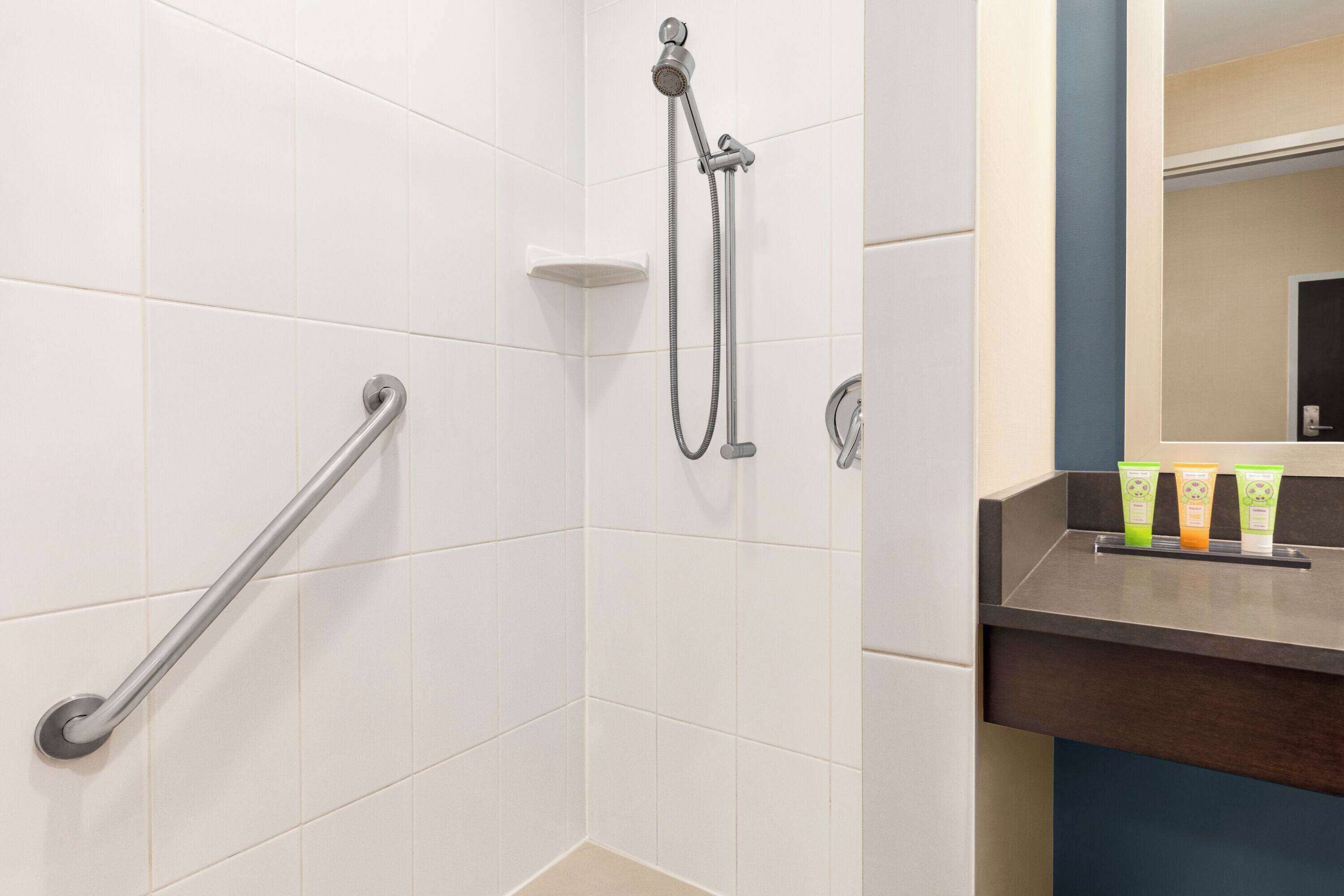 Une salle de bain carrelée de carreaux blancs, une douche avec un pommeau de douche amovible, une barre d'appui et un meuble-lavabo avec des articles de toilette.