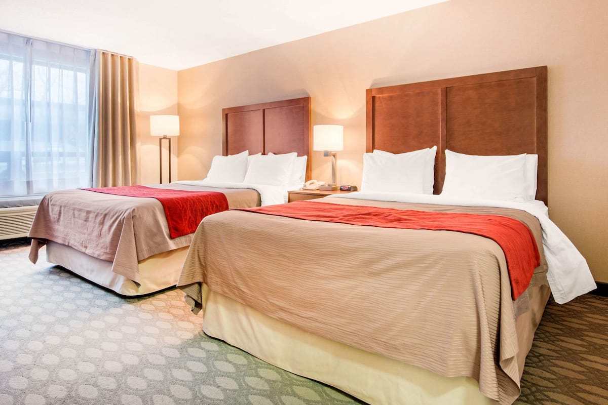 A hotel room with two beds, each with a wooden headboard, a red accent throw, and a bedside table with a lamp.