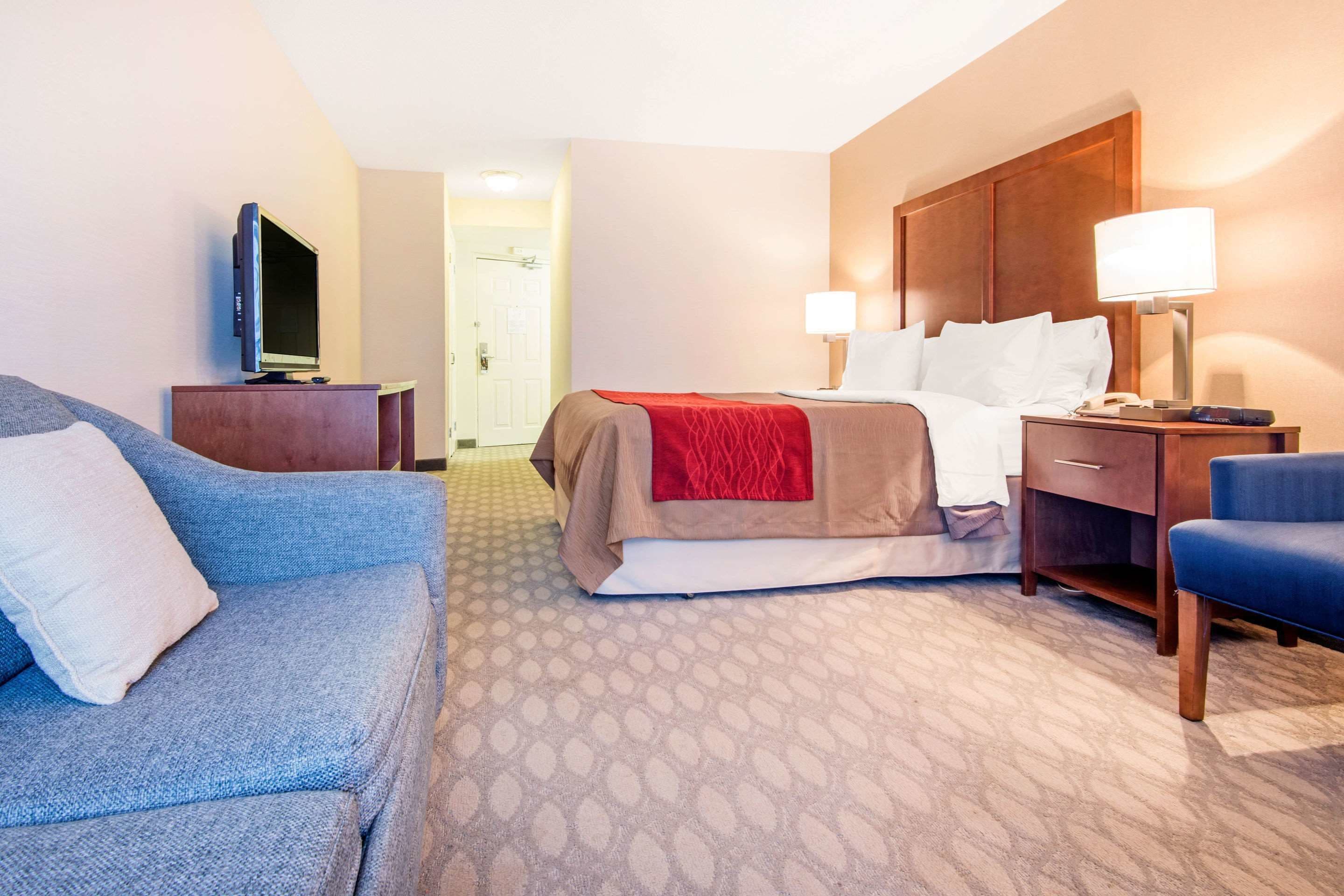 A hotel room with a bed, a TV on a stand, a desk with a lamp, and a blue armchair.