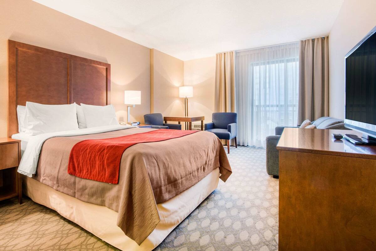 A hotel room with a large bed, a desk, two chairs, a TV, and a window with curtains.
