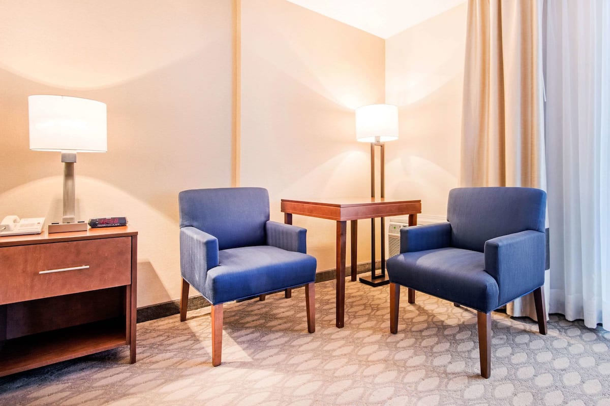 A hotel room with two blue armchairs, a small wooden table, a desk with a lamp, and a telephone.
