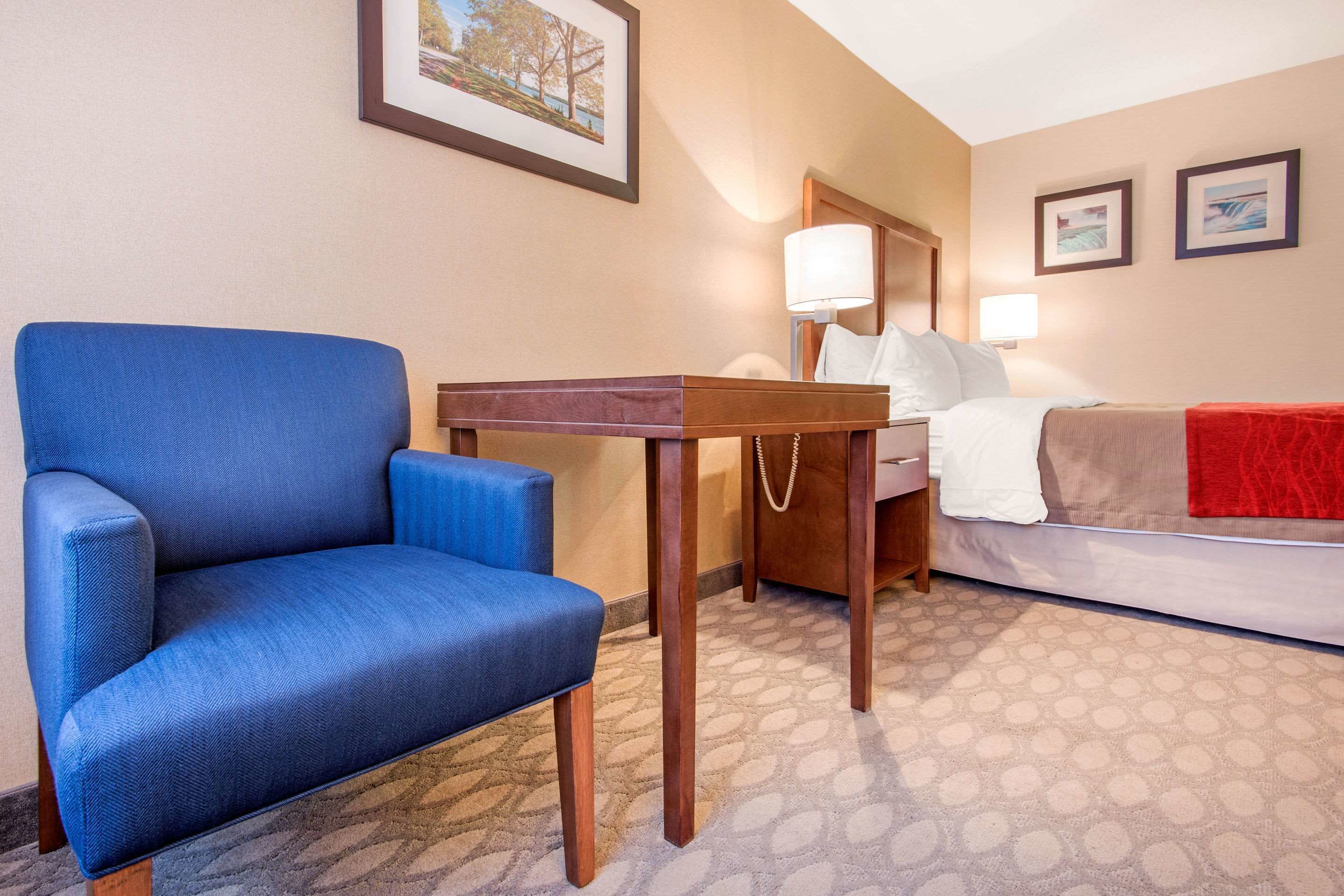 A hotel room with a blue armchair, a wooden nightstand, a bed with a red blanket, and two wall-mounted lamps.