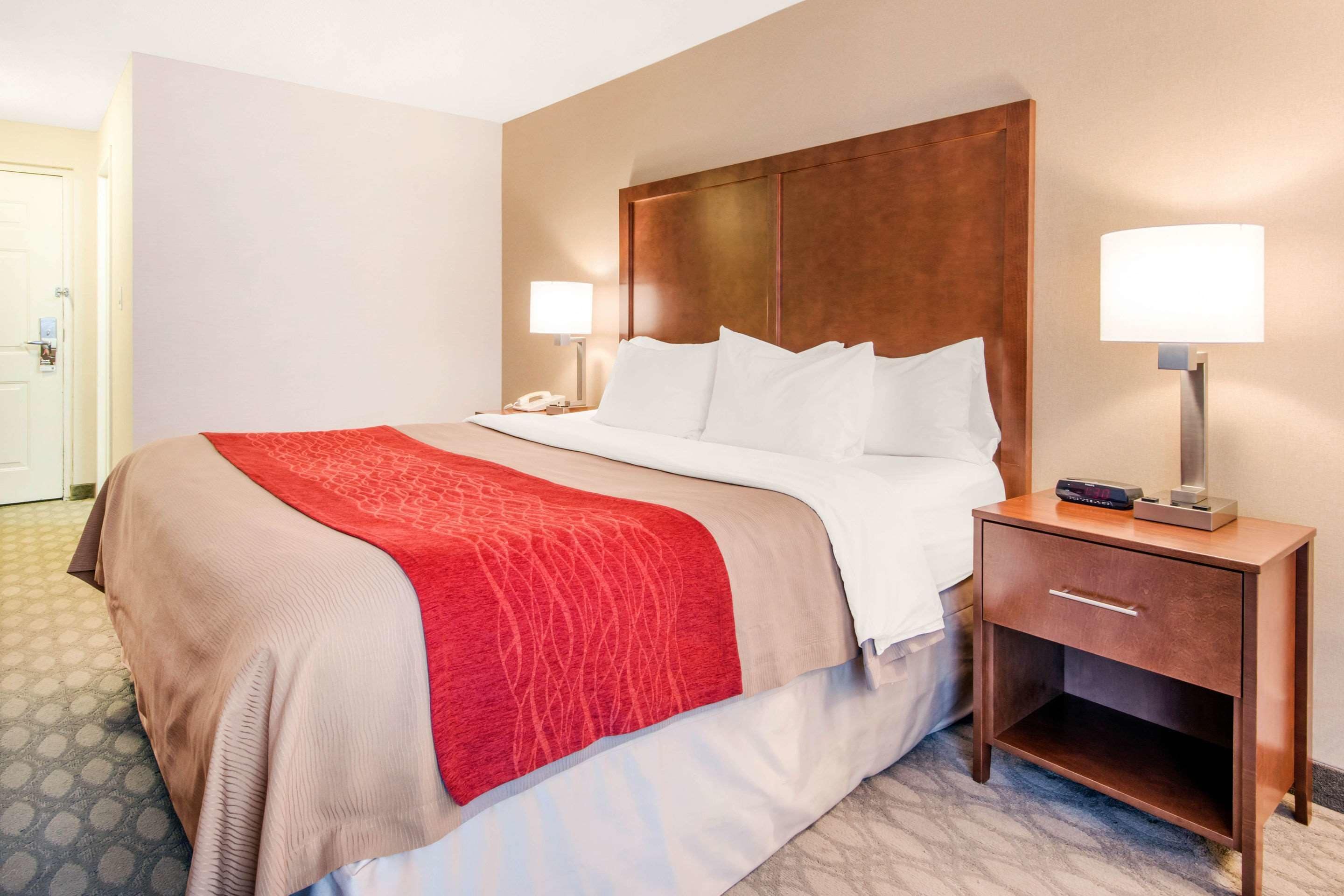 A hotel room with a large bed, a wooden headboard, two bedside lamps, and a nightstand with a phone and a remote control.