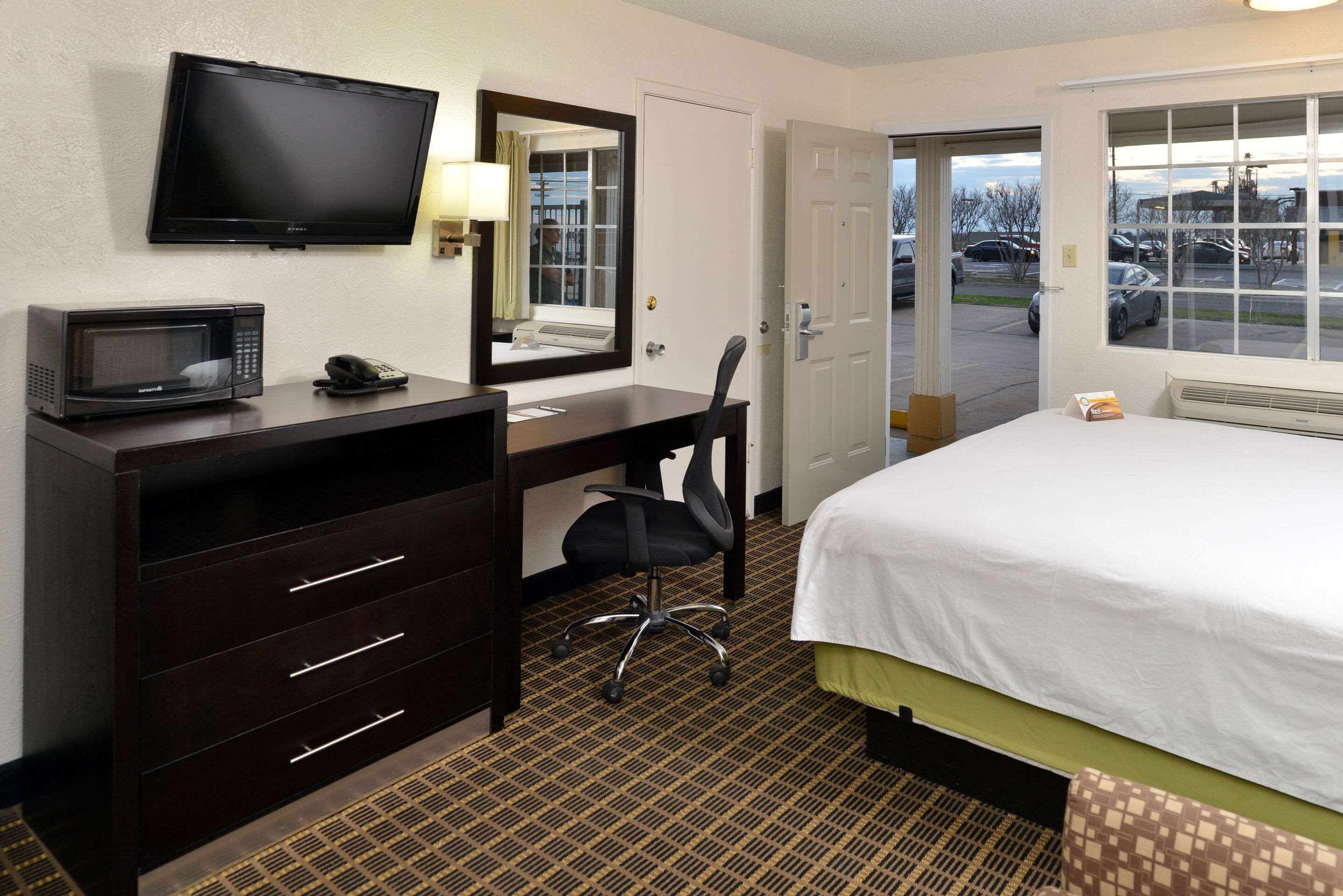 Ein Hotelzimmer mit Bett, Schreibtisch mit Stuhl, Mikrowelle, Telefon, Fernseher und Fenster mit Blick auf geparkte Autos und Gebäude.