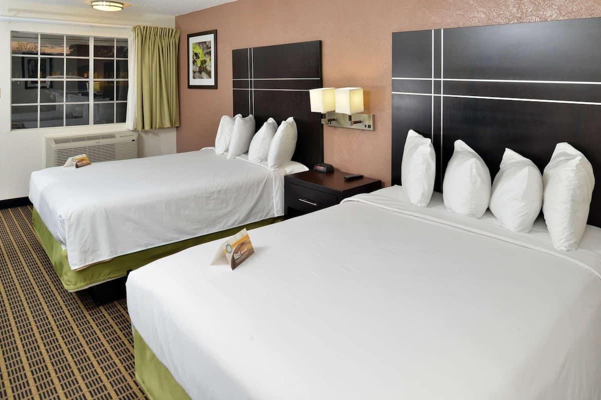 A hotel room with two beds, a nightstand with a lamp, a window with curtains, and a wall-mounted artwork.