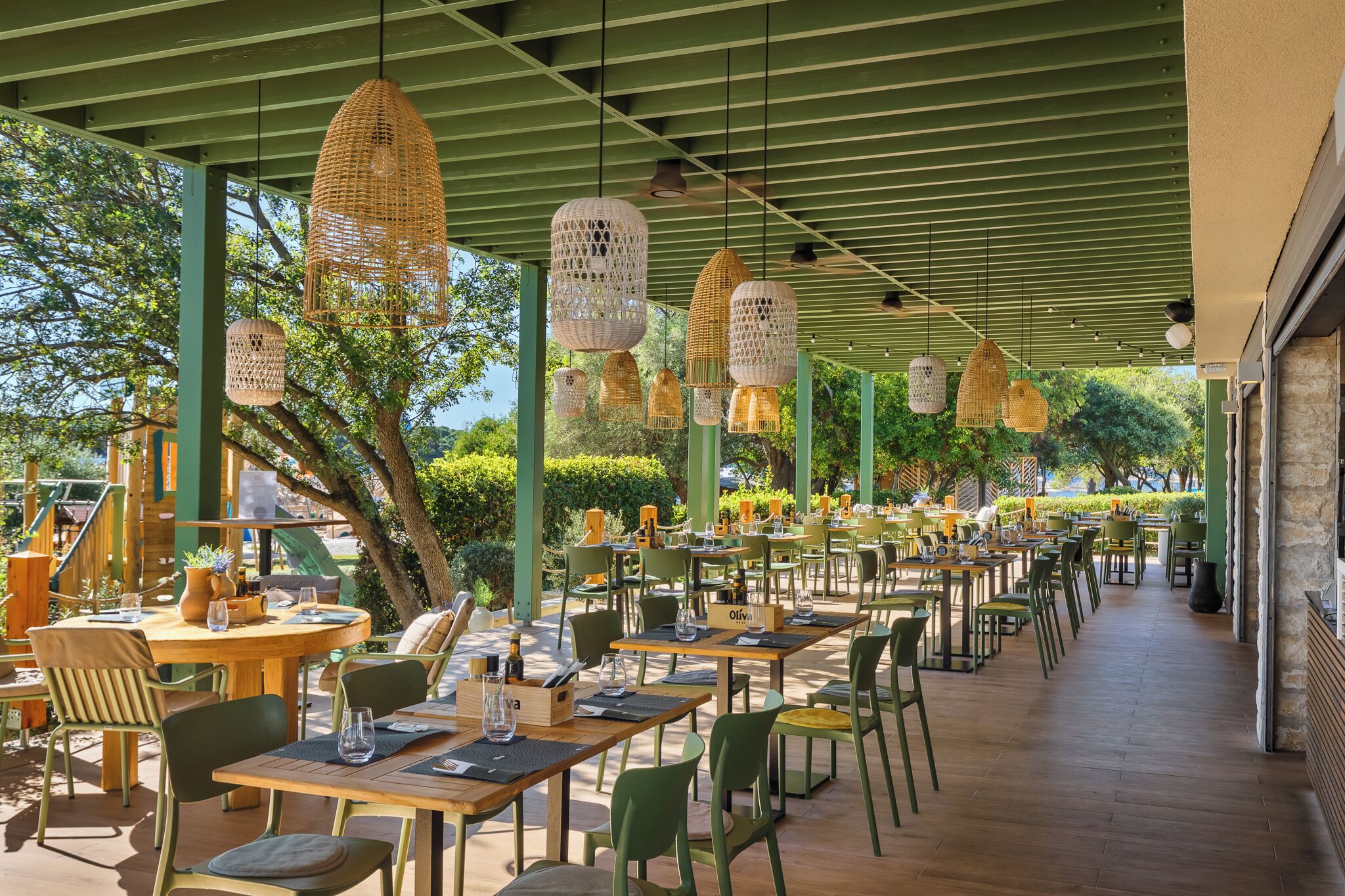An outdoor dining area with tables and chairs, hanging wicker lamps, and a green roof structure.