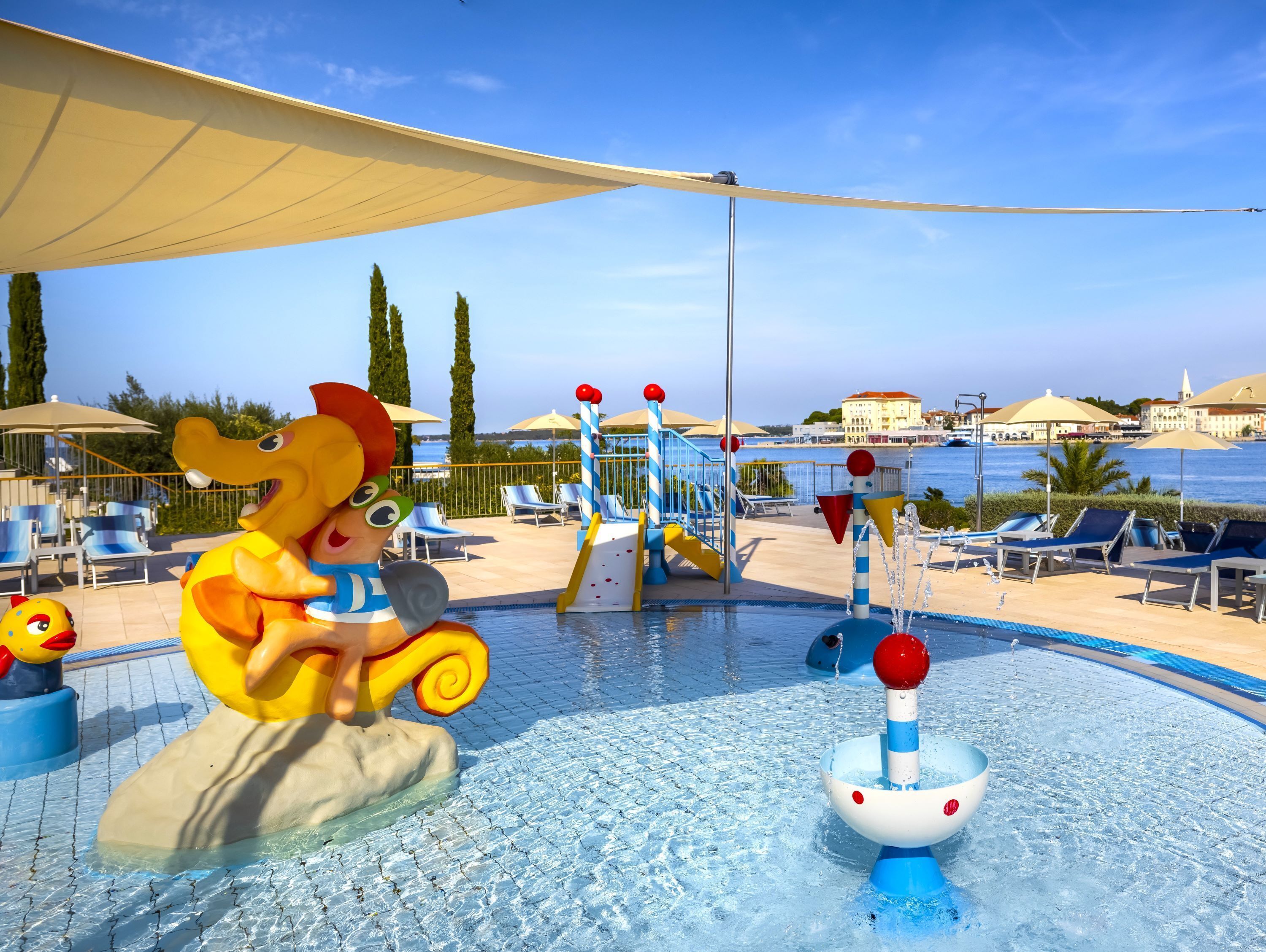 A pool area with a water play structure, lounge chairs, and a view of the sea.