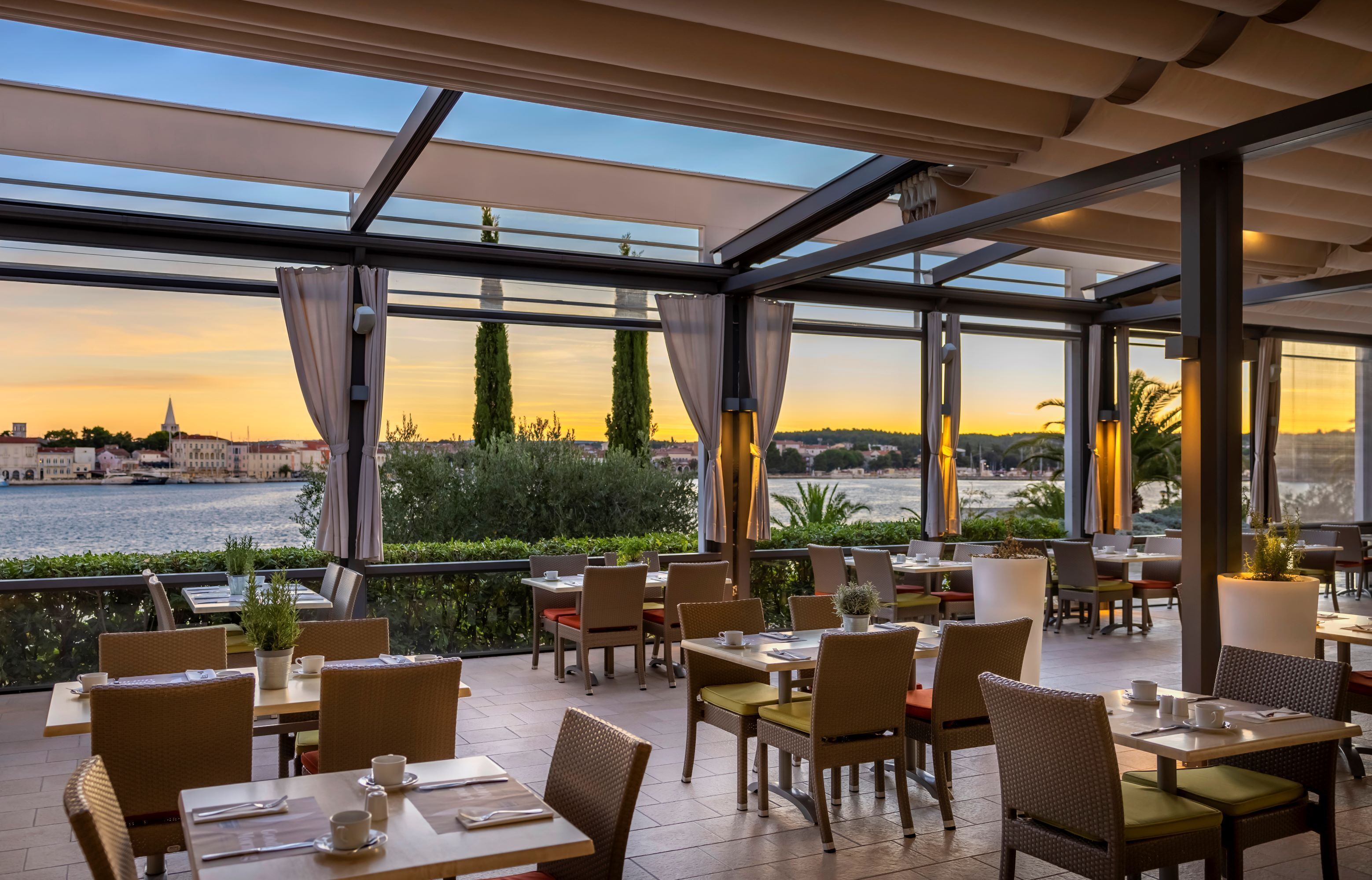 An outdoor dining area with tables and chairs, overlooking a river and cityscape at sunset.