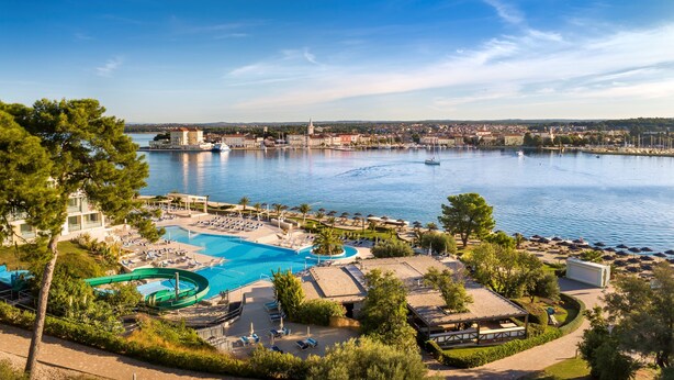 A resort with a large swimming pool, a water slide, and a view of the city across the water. - Isabella Island Hotel & Villas, Valamar Collection (Porec)