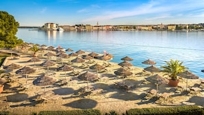 A beach with sun loungers and thatched umbrellas, a clear view of the water, and a distant cityscape. - Isabella Island Hotel & Villas, Valamar Collection (Porec)