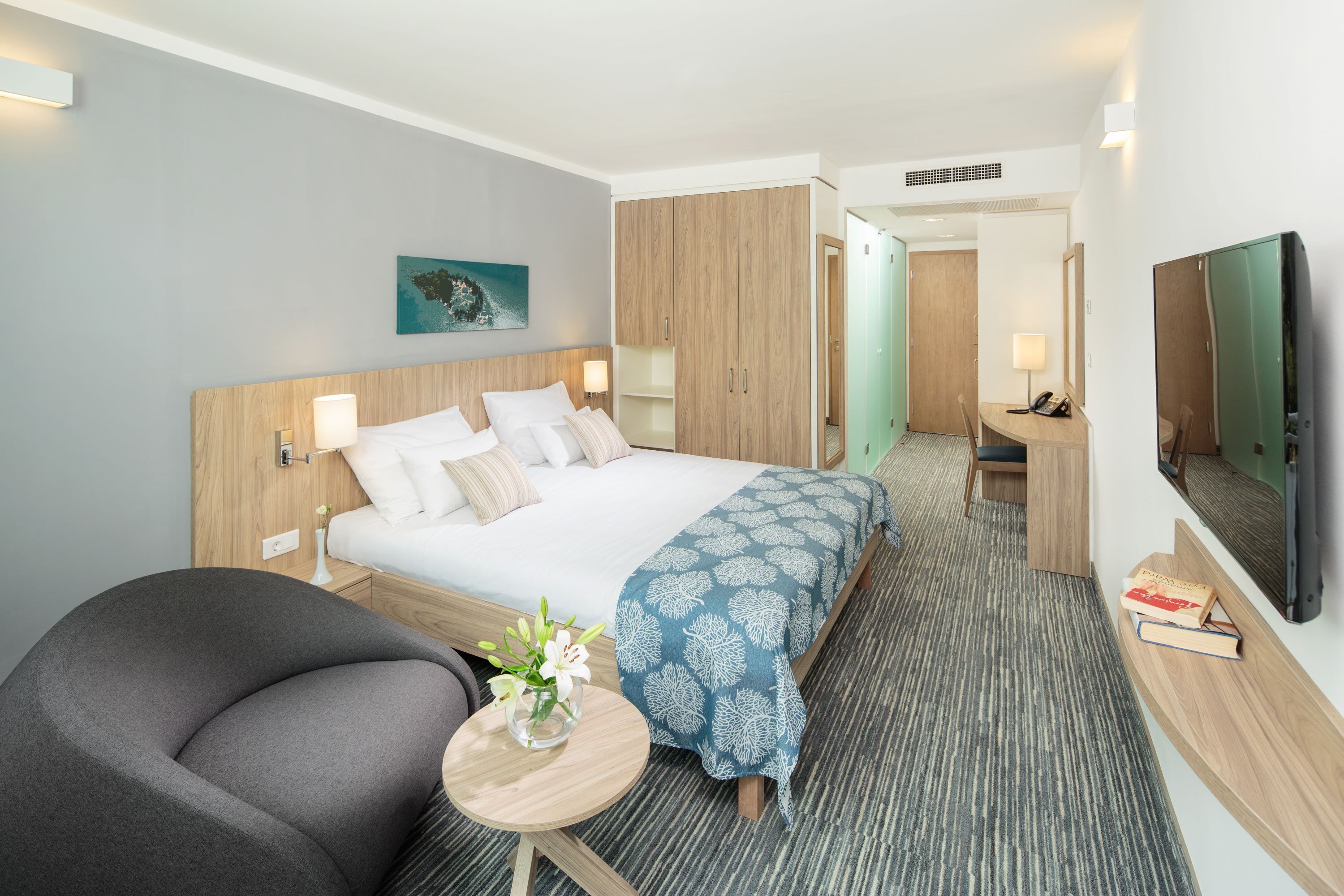 A hotel room with a bed, a desk, a chair, a TV, and a small table with a vase of flowers.