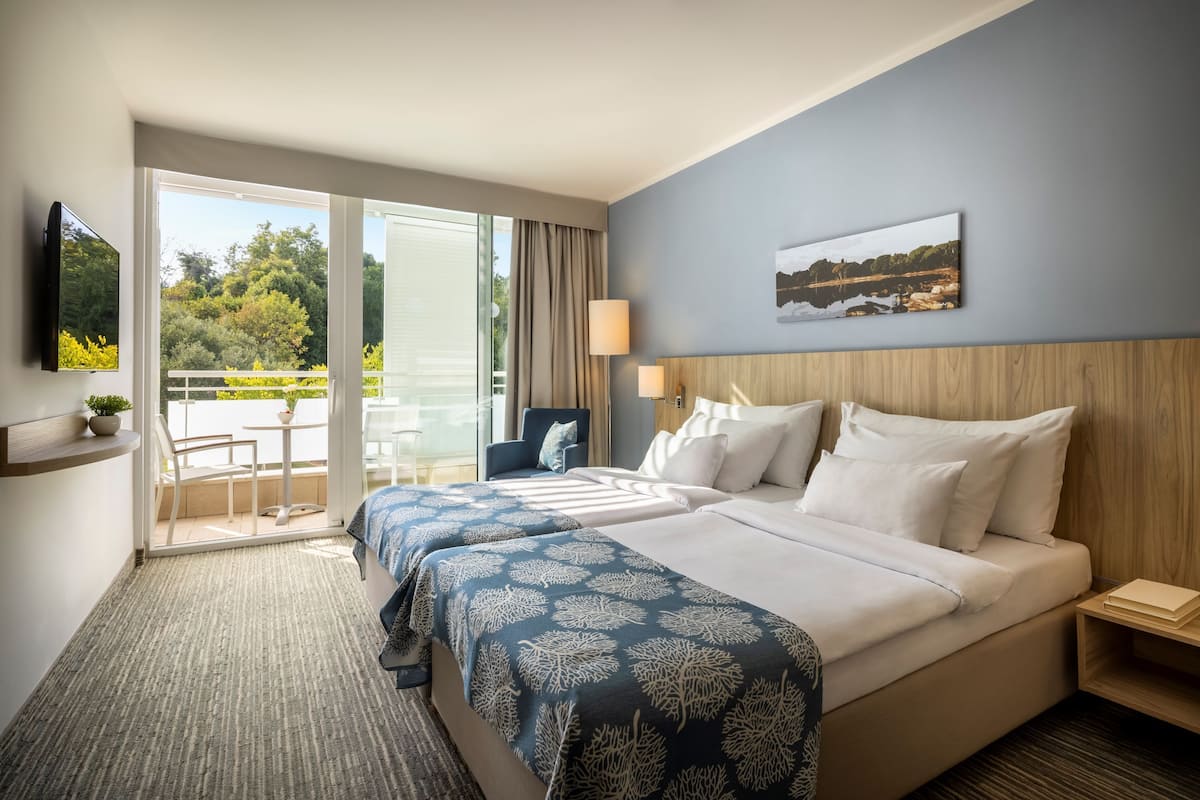 Una camera d'albergo con due letti, un balcone vista alberi, una TV a parete e un quadro appeso.