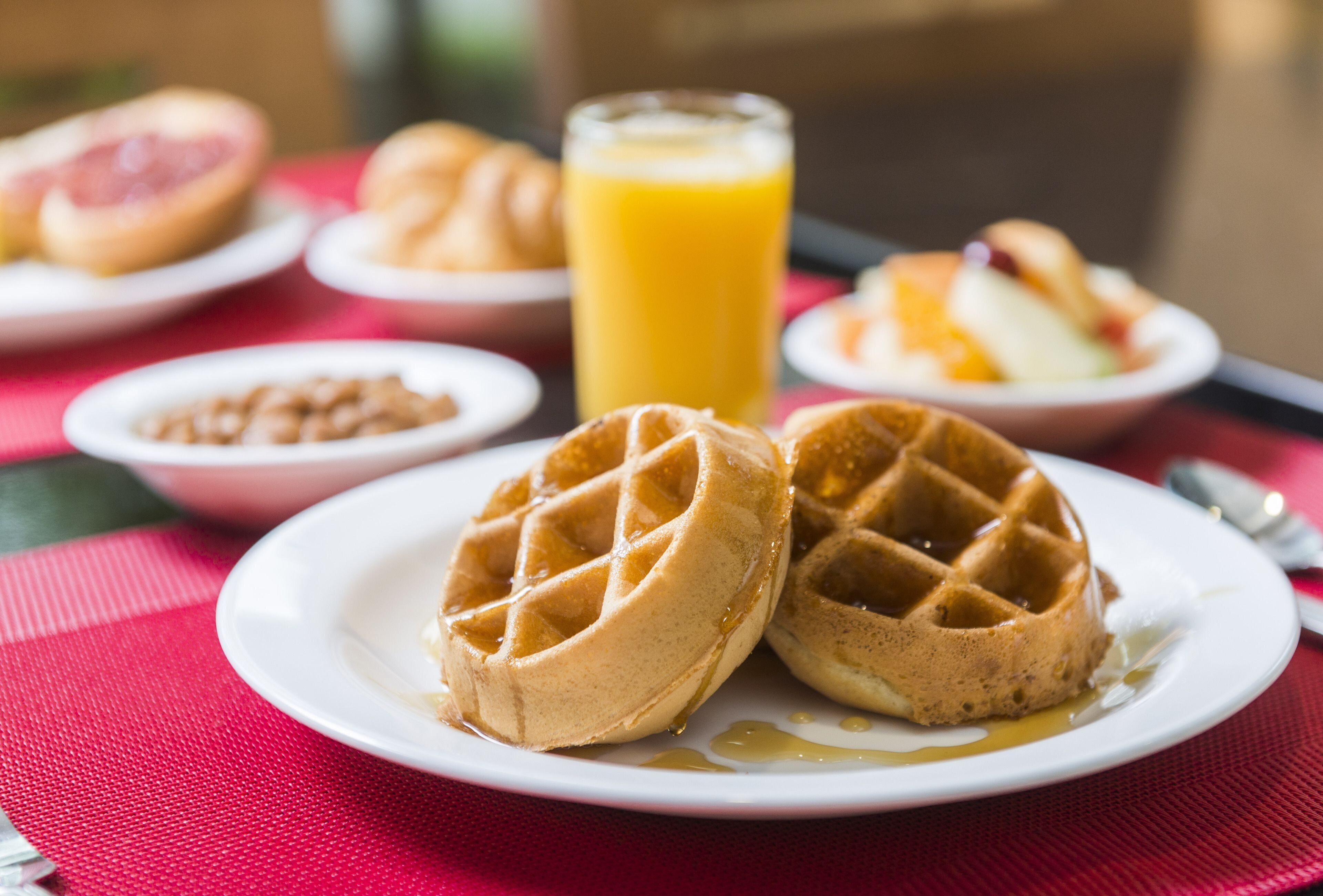 Un desayuno con waffles, un vaso de zumo de naranja y diversas frutas en platos.