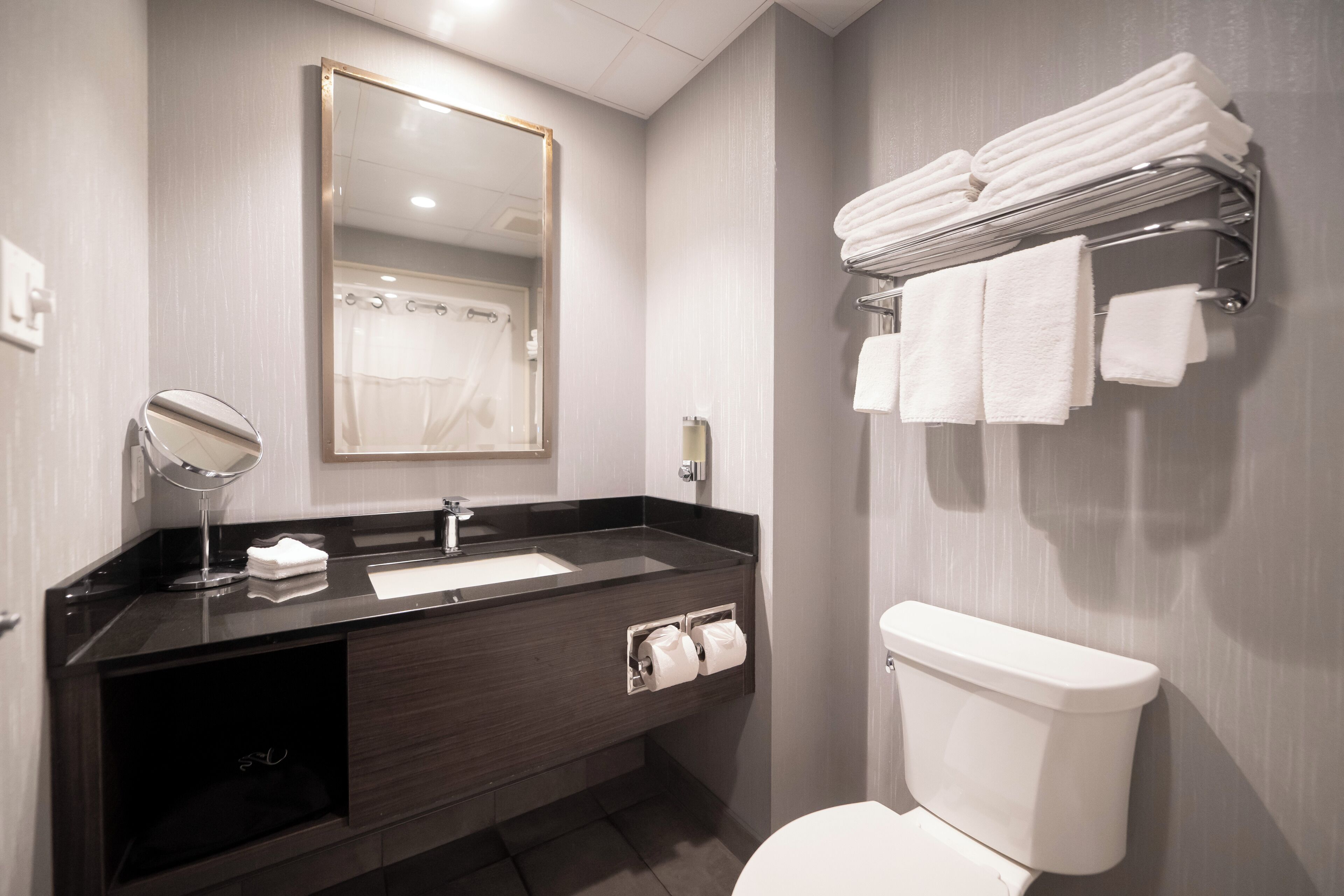 Une salle de bain moderne avec un meuble-lavabo en bois foncé, un grand miroir, des toilettes blanches et un porte-serviettes où sont soigneusement pliées les serviettes.