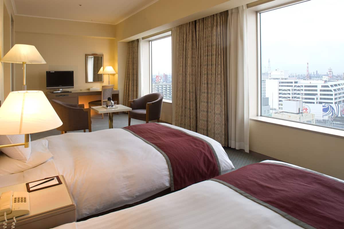 Hotelkamer met twee bedden, een bureau met een televisie, een stoel en uitzicht op de stad.