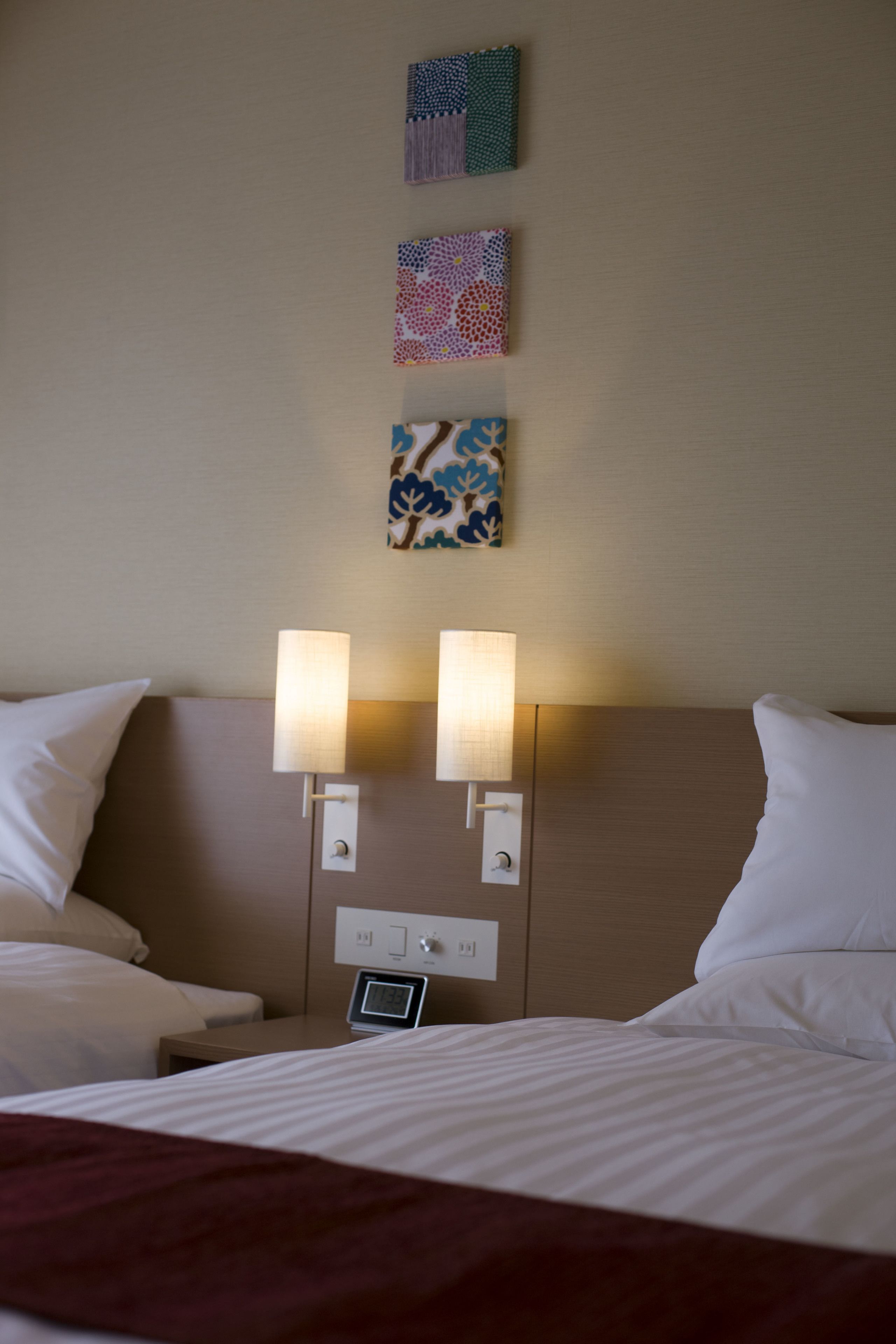 A hotel room with a bed, two bedside lamps, a digital clock, and three patterned wall decorations.
