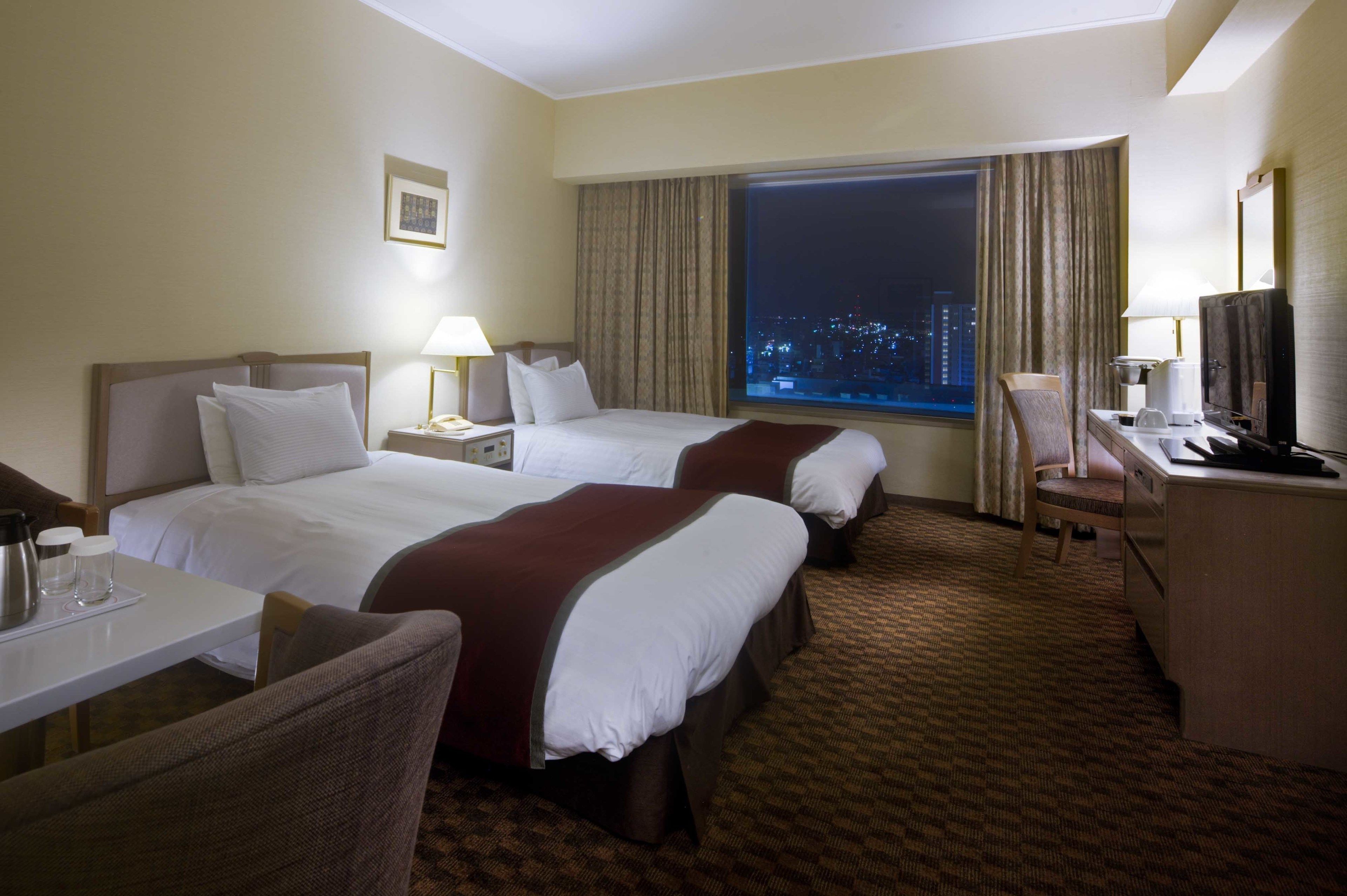 A hotel room with two beds, a desk, a chair, a television, and a large window with a city view at night.