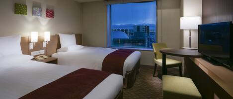 A hotel room with two beds, a desk with a television, a chair, and a view of a cityscape at dusk.