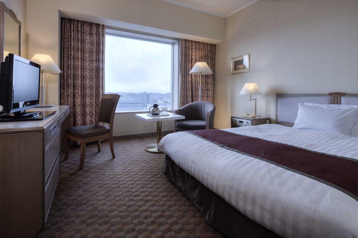Een hotelkamer met een bed, een bureau met een televisie, een stoel, een klein tafeltje met een koffiezetapparaat, en een raam met gordijnen.