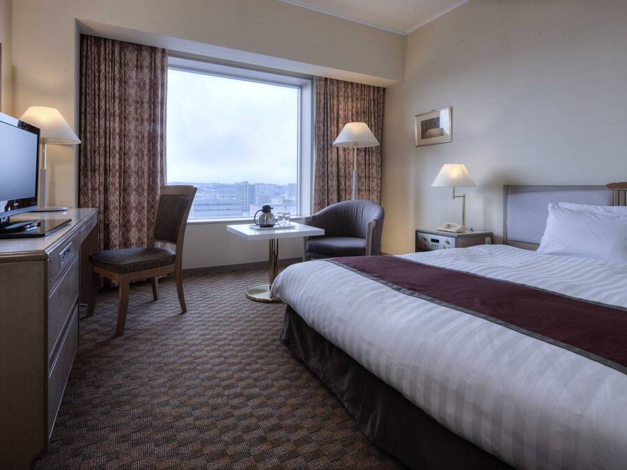 A hotel room with a bed, a desk with a television, a chair, a small table with a coffee pot, and a window with curtains.