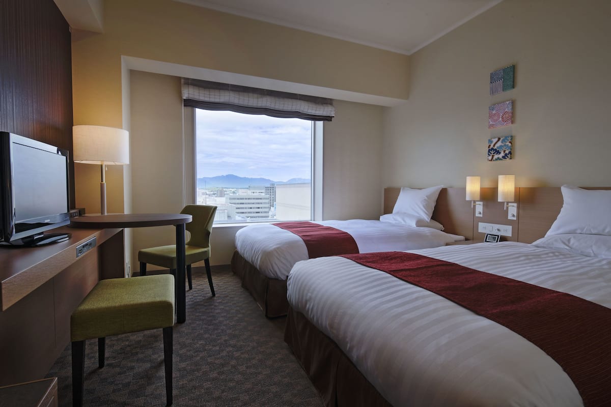 Een hotelkamer met twee bedden, een bureau met stoel, een televisietoestel en een raam met uitzicht op de stad.